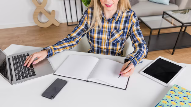 A girl sits at a table and writes something, next to her is a laptop and a phone