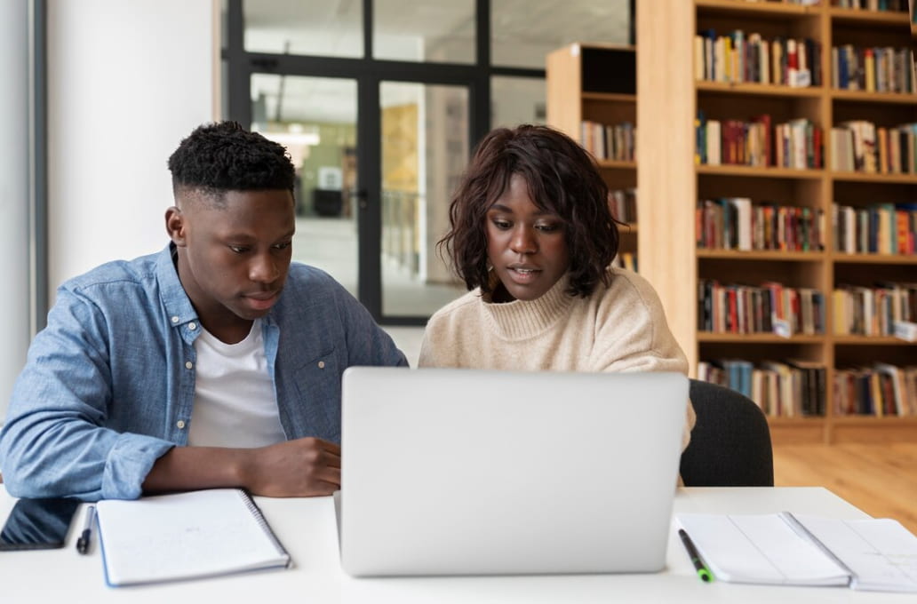 Two students looking at the opened silver laptop in a library, the notes and pen on the table